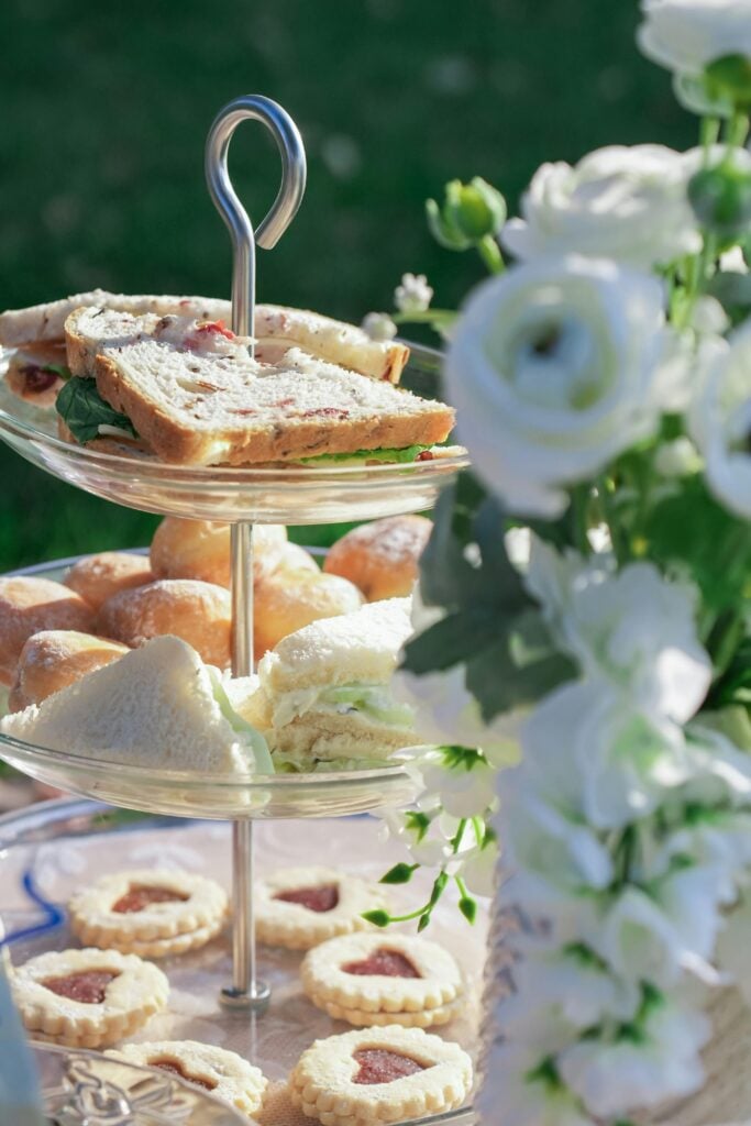 A three-tiered glass stand with cookies, tea sandwiches, and scones on a table outside with white flowers as a bridal shower tea party idea.