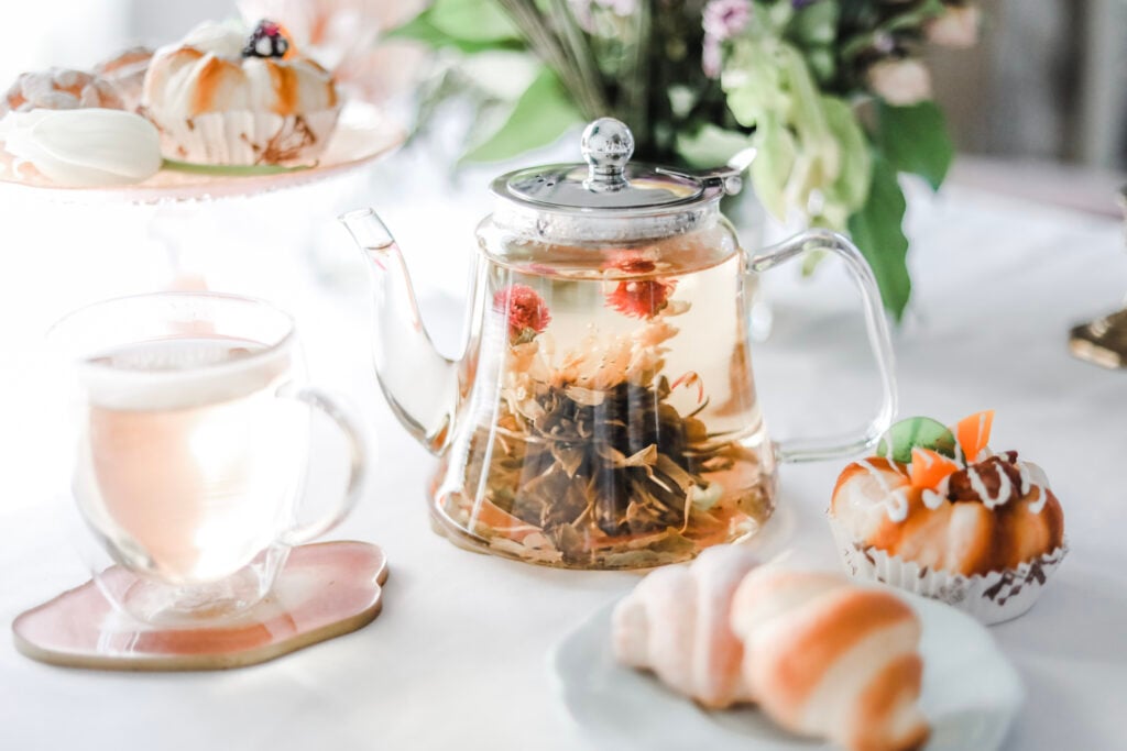 Glass teapot with blooming tea, a teacup, and pastries on a white table.