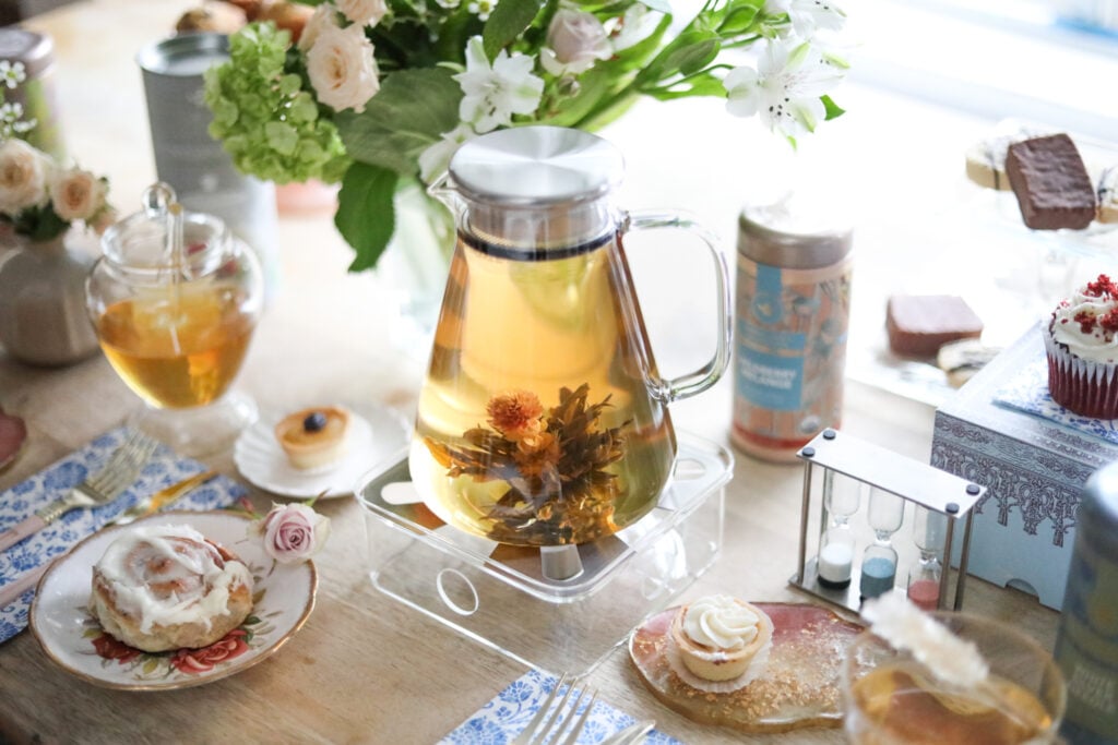 A round glass teapot on a glass warmer on a table with fresh flowers, sweets, and tea accessories for tea connoisseurs.