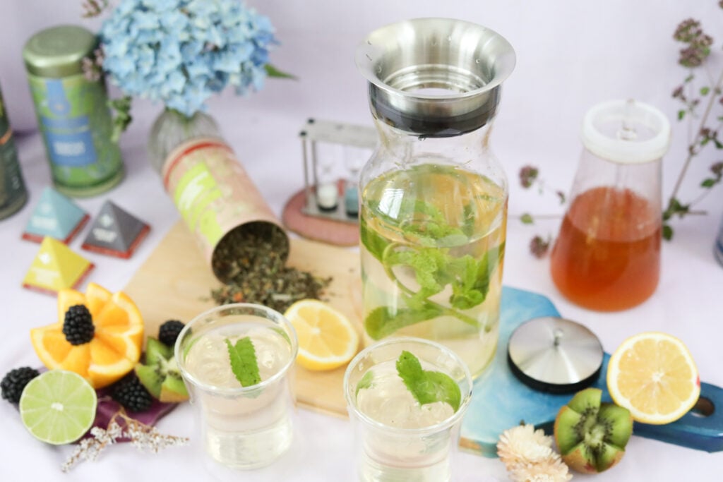 A carafe of infused tea with mint leaves on a table with two glasses and fresh ingredients.