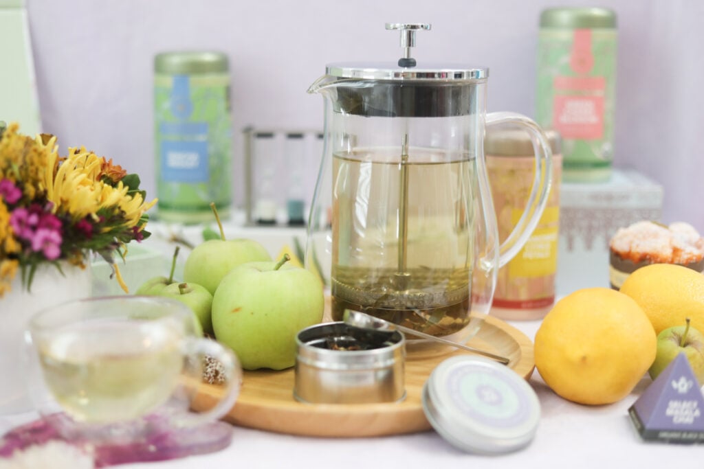 Glass tea press and teaware with green tea surrounded by fresh apples, lemons, tea, and flowers to help with green tea health benefits.