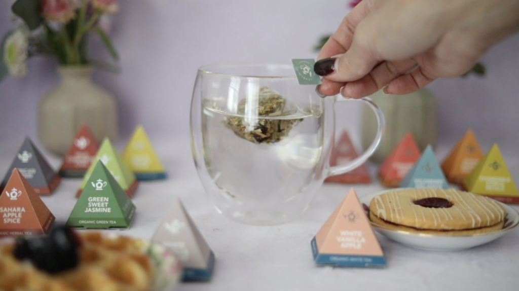 Someone’s hand holding a Teabloom tea sachet tag while steeping a glass cup of tea surrounded by tea sachet pyramid packages.