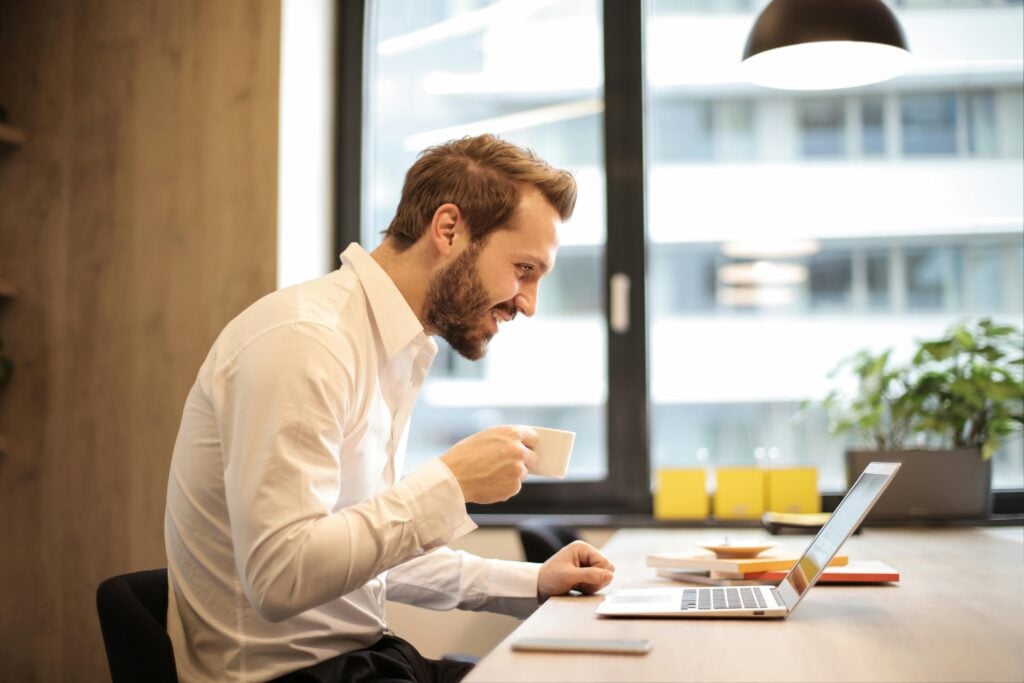 A man in the office smiling while looking at his laptop and holding a cup of green tea with many health benefits.