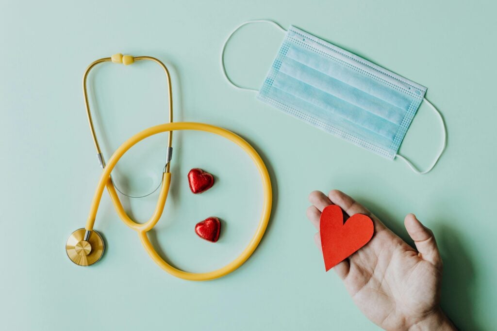 A hand holding a paper red heart next to a face mask, a yellow stethoscope, and two small red foil hearts on a light blue table.