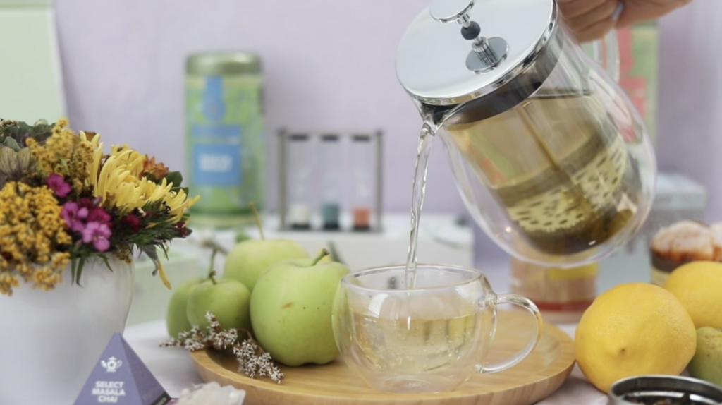 Someone pouring green tea from a tea press into a glass teacup next to fresh apples, lemons, and flowers.