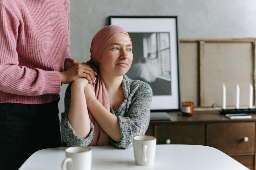 A person consoling a young woman with a chemotherapy head wrap sitting at a table with two cups of green tea for health benefits.
