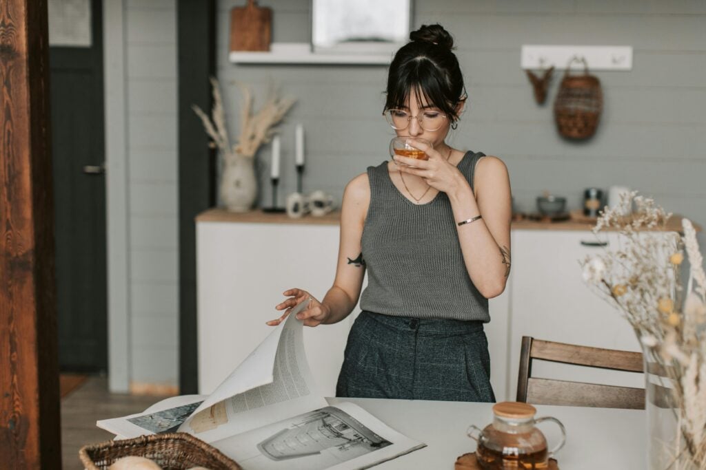 A woman with a grey shirt and pants standing at a kitchen table with a magazine sipping on a cup of tea with a teapot on the table.
