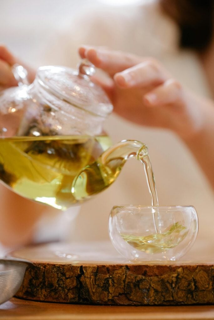 A woman pouring a teapot of green tea for health benefits into a glass teacup.