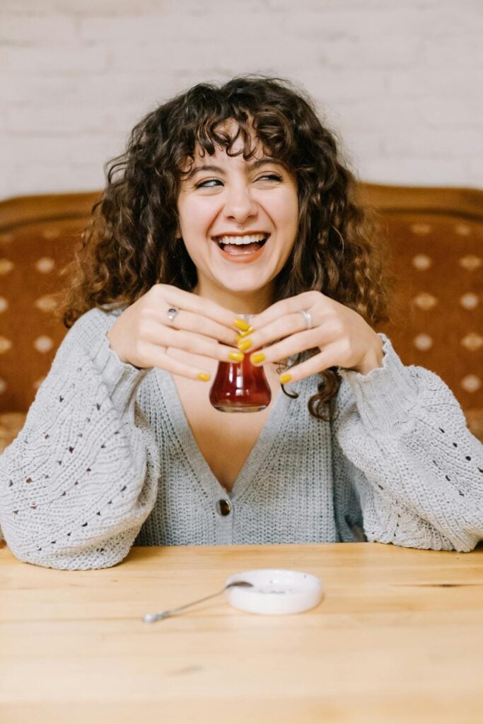 Woman laughing and smiling wearing a grey sweater and holding a tulip-shaped unique tea cup full of Maghrebi tea.
