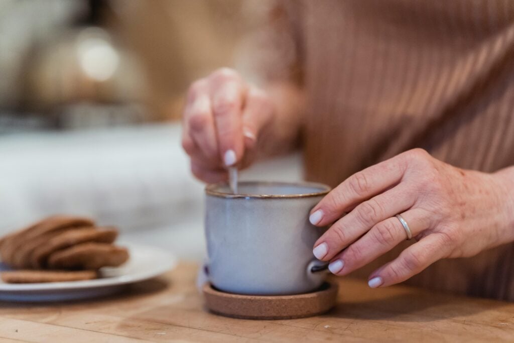 Closeup of a woman stirring a mug of hot tea with interesting tea flavors and with a plate of snacks on a wooden counter.