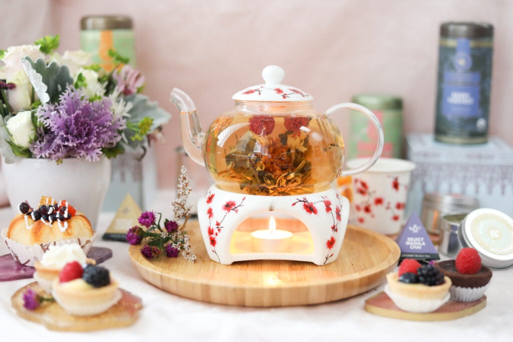 A beautiful glass and porcelain blooming flower tea set with red cherry blossoms on a table surrounded with fresh flowers, pastries, and canisters of loose-leaf teas from Teabloom.