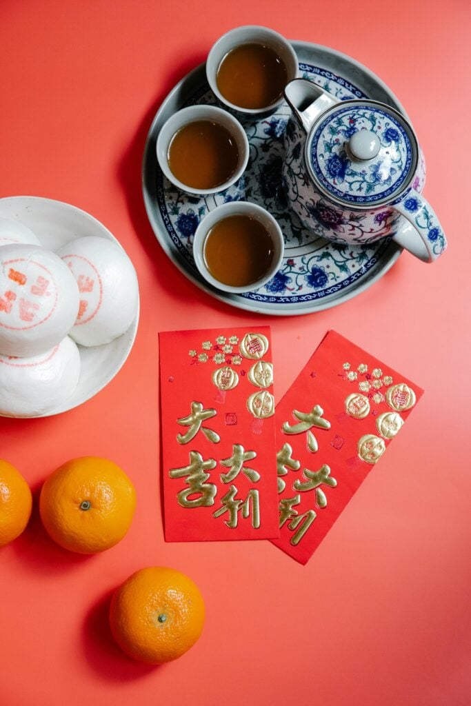 Red and gold Chinese New Year envelopes on a red table with mandarins, Chinese snacks, and a blue and white tea set.