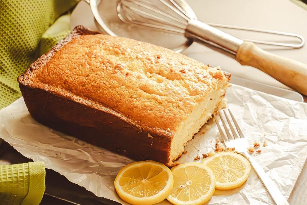 A loaf of lemon drizzle cake next to lemon slices and kitchen utensils. 
