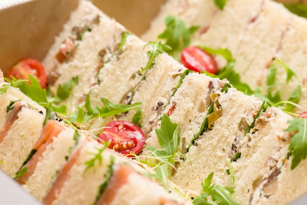 Finger sandwiches cut into triangles and displayed elegantly on a table with a sprinkle of tomatoes and arugula. 