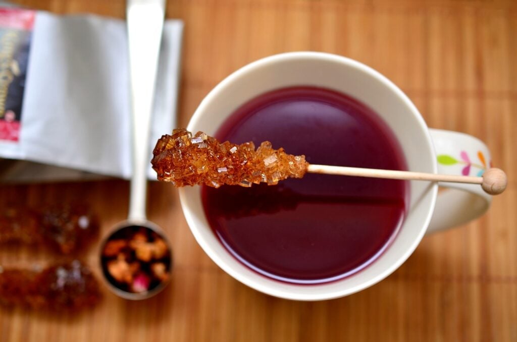 A top-down view of black tea in a white teacup with a stick of sugar and a spoon of loose leaf tea sitting on a table.