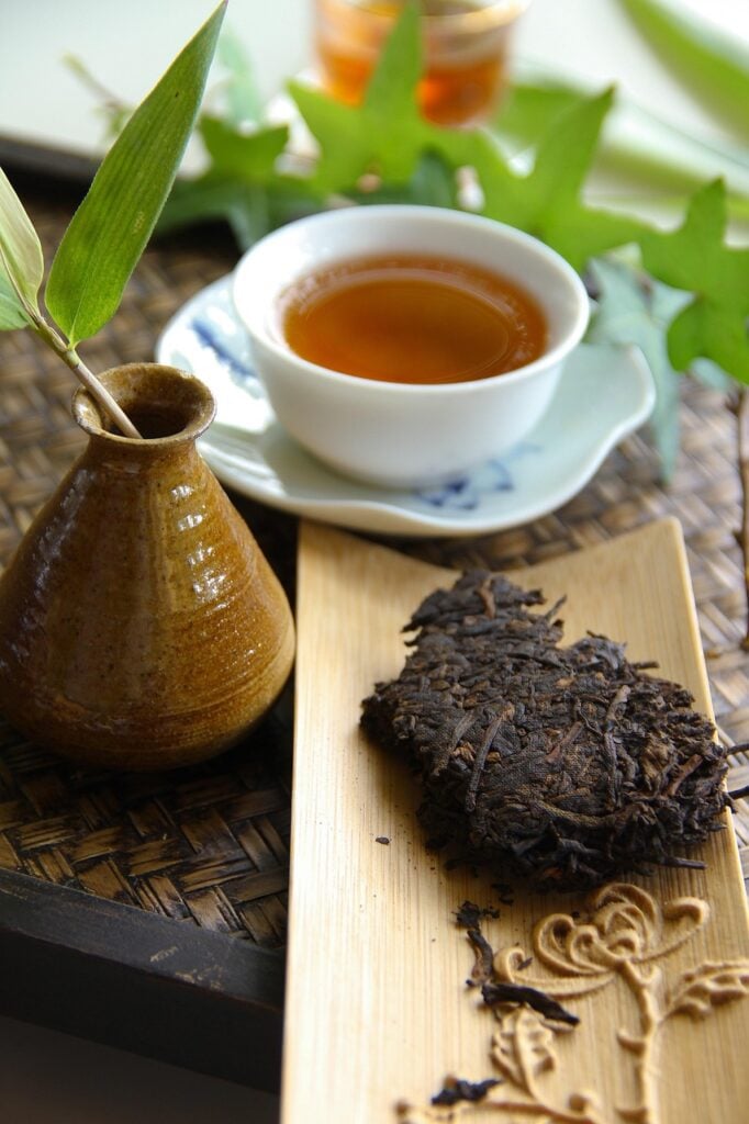 A broken block of pu-erh tea next to a steeped cup of tea and greenery displayed as an example of an aged tea from around the world.