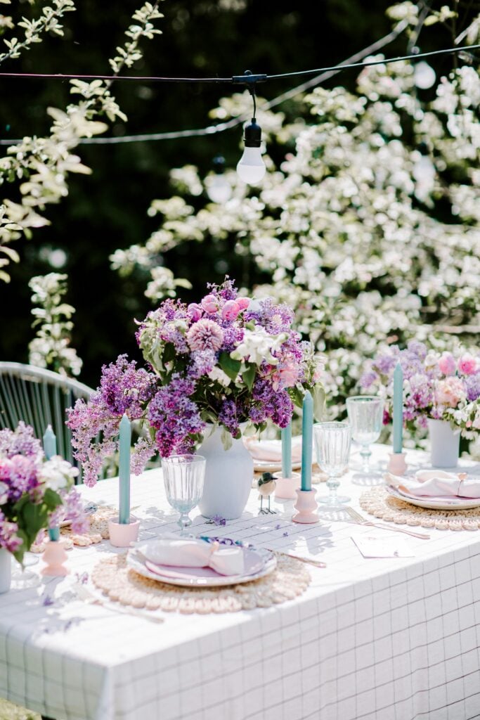 A garden tea party table with white and pink accents surrounded by purple and white flowers with tall blue candles.
