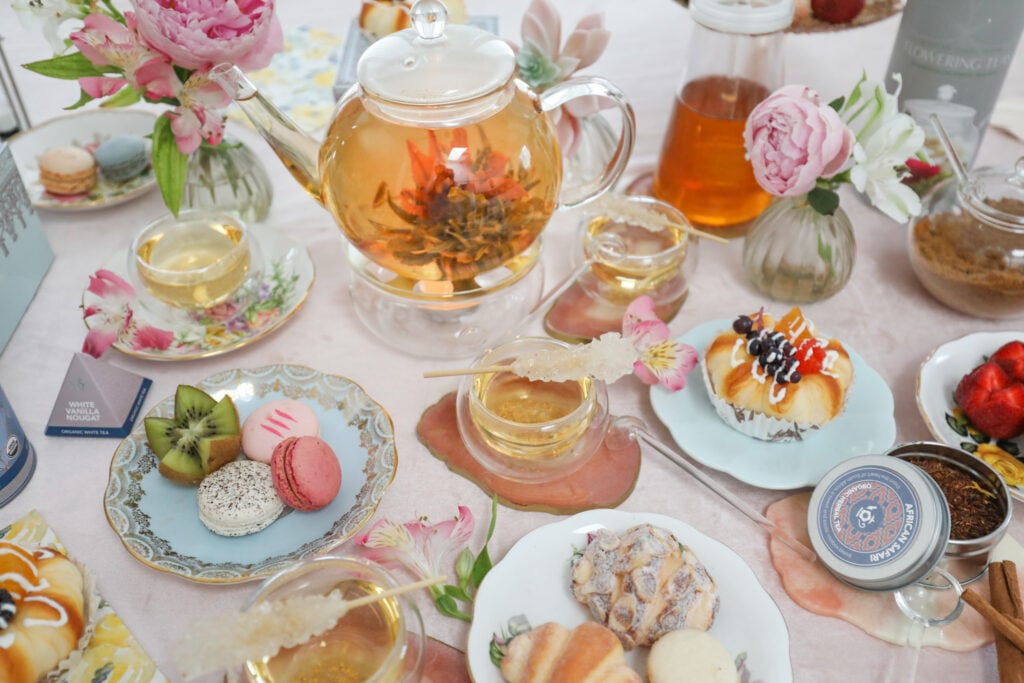 Teabloom glass teaware display with teacups and teapot on a white table with pastries, flowers, and garden tea party treats.