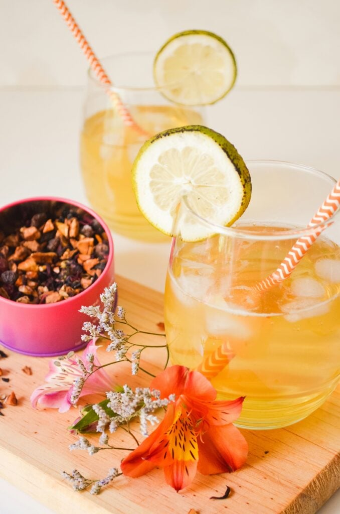 Glasses of iced tea with lime slices on a cutting board with loose-leaf tea and fresh-cut orange flowers.