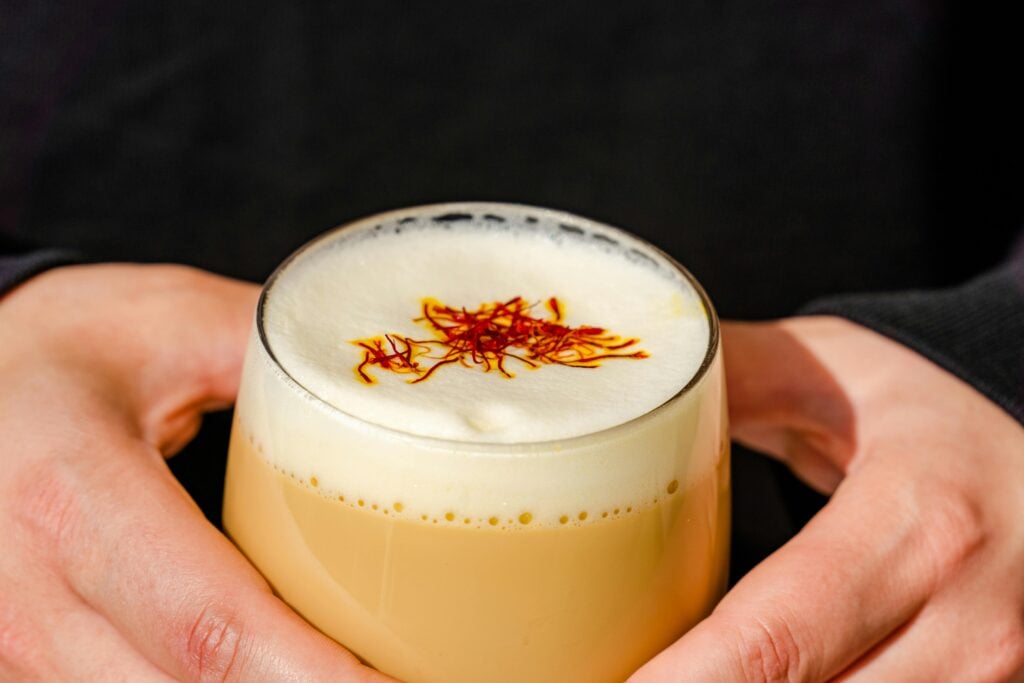 A person holding a glass cup full of milky, foamy, Teh Tarik sprinkled with saffron.