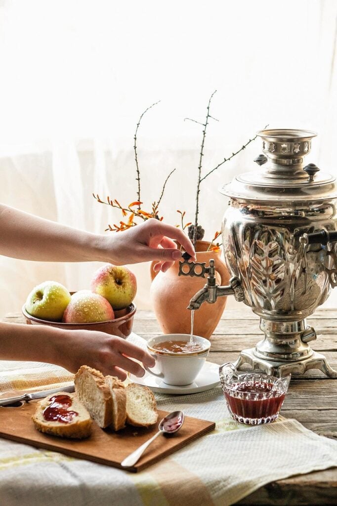 A table with bread, jam, fruit, and a Russian samovar with a woman pouring a serving of tea from around the world into a teacup.