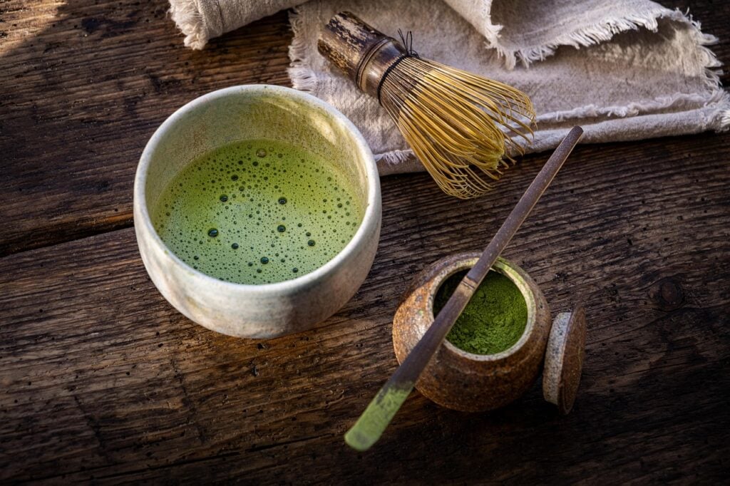 A traditional cup of whisked bright green matcha tea with a bamboo whisk and tea powder container on a wooden table.
