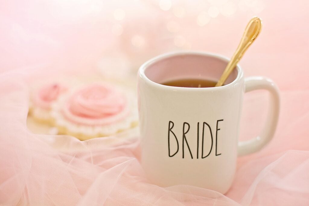 A white mug with the word BRIDE filled with hot tea and a gold spoon on a pink chiffon tablecloth with pink frosting pastries.