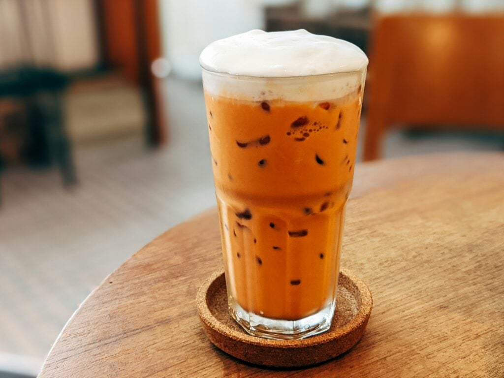 A white and orange Thai iced tea on a cork coaster sitting on a wooden table.
