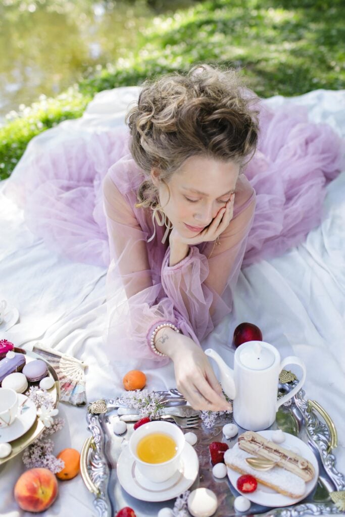 A woman in a flowy purple dress on the grass with a short table full of garden tea party pastries, a teacup, and fruit.