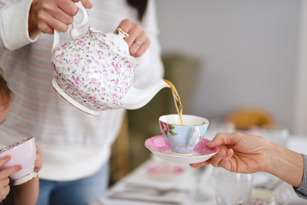 A woman pouring a pot of tea into a teacup for a garden tea party guest.