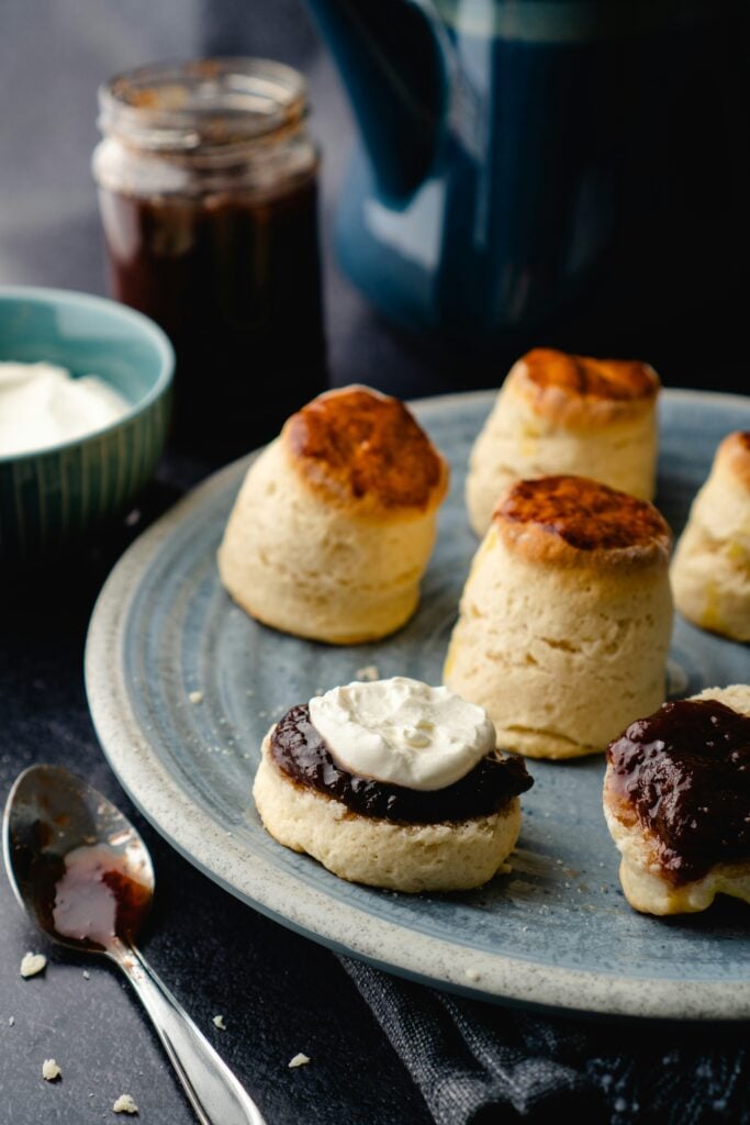 A plate of scones with one scone cut in half with jam and clotted cream on top.