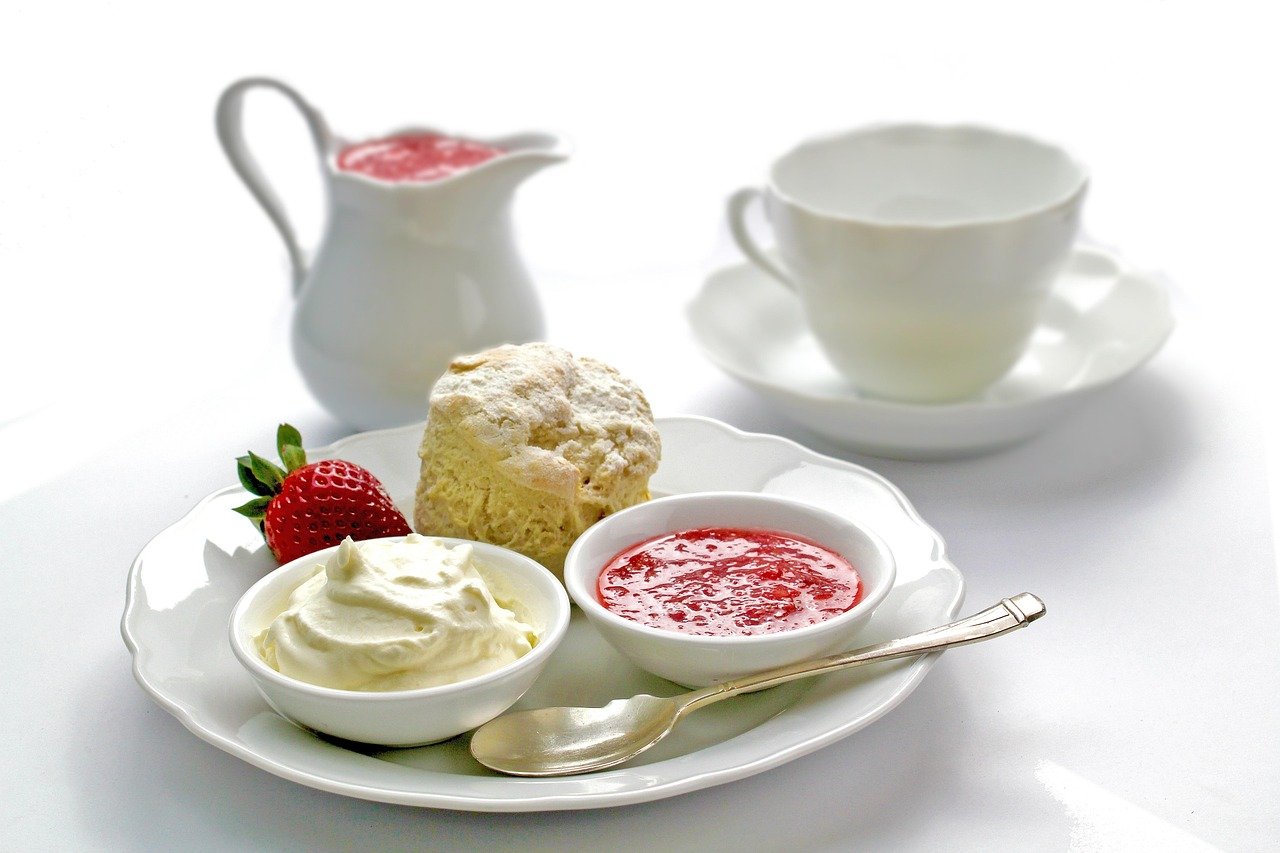 A white cup and saucer, pitcher, and plate with a scone, strawberry, cream, and strawberry jam with a silver spoon.