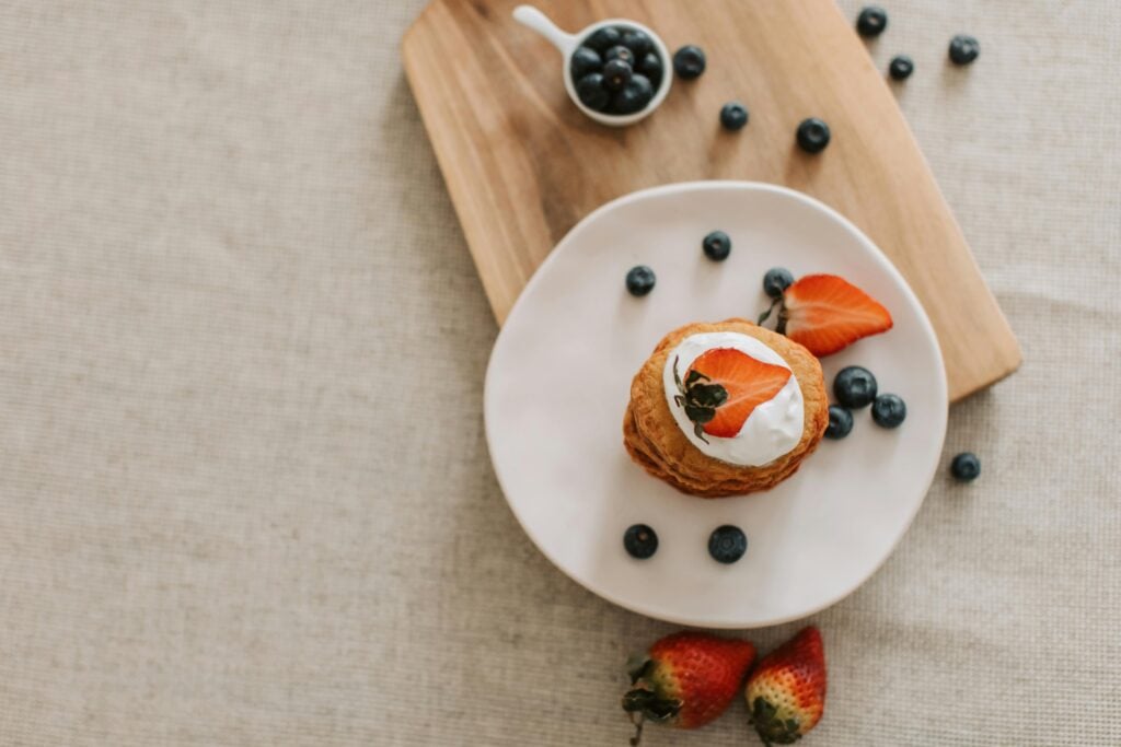 A wooden tray with scattered blueberries and a white plate with a scone, cream, and fresh strawberries.
