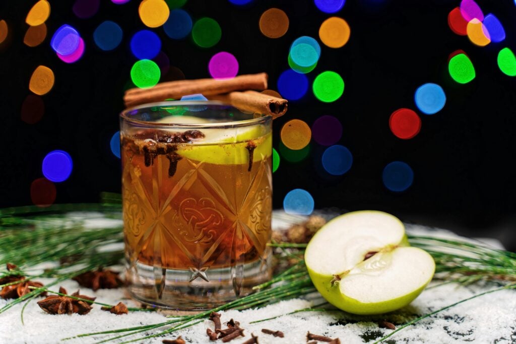 Cinnamon apple tea cocktail recipe with fresh apple slices on a table with spices and star anise with colorful holiday lights in the background.