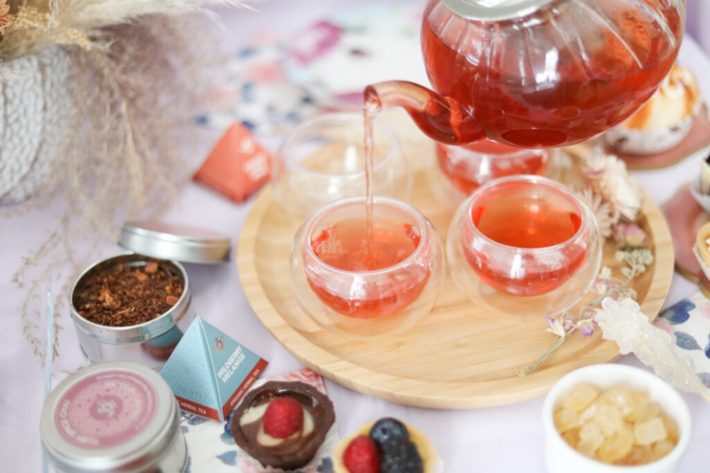 Teabloom glass teapot pouring black tea into round teacups with sweets on the side