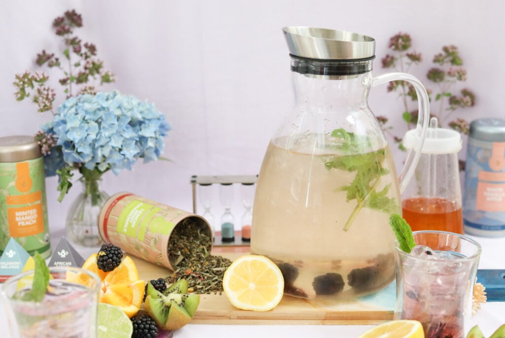 Borosilicate glass iced tea pitcher with herbs and berries, surrounded by flowers, fruit, and green tea.