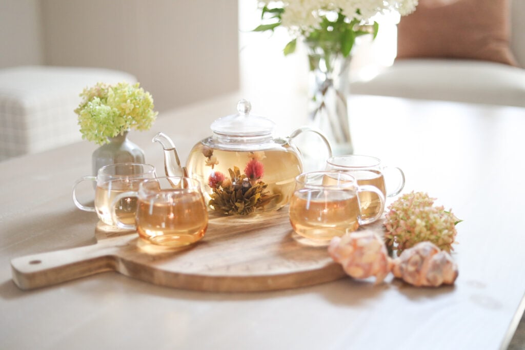 A glass teapot with a blooming tea flower and four teacups with tea on a wooden board displayed on a table with vases of flowers and two small pastries.