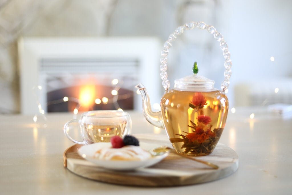 Glass teapot with blooming tea and a glass teacup on a wooden charger with a pastry on a white plate in front of a white living room fireplace.