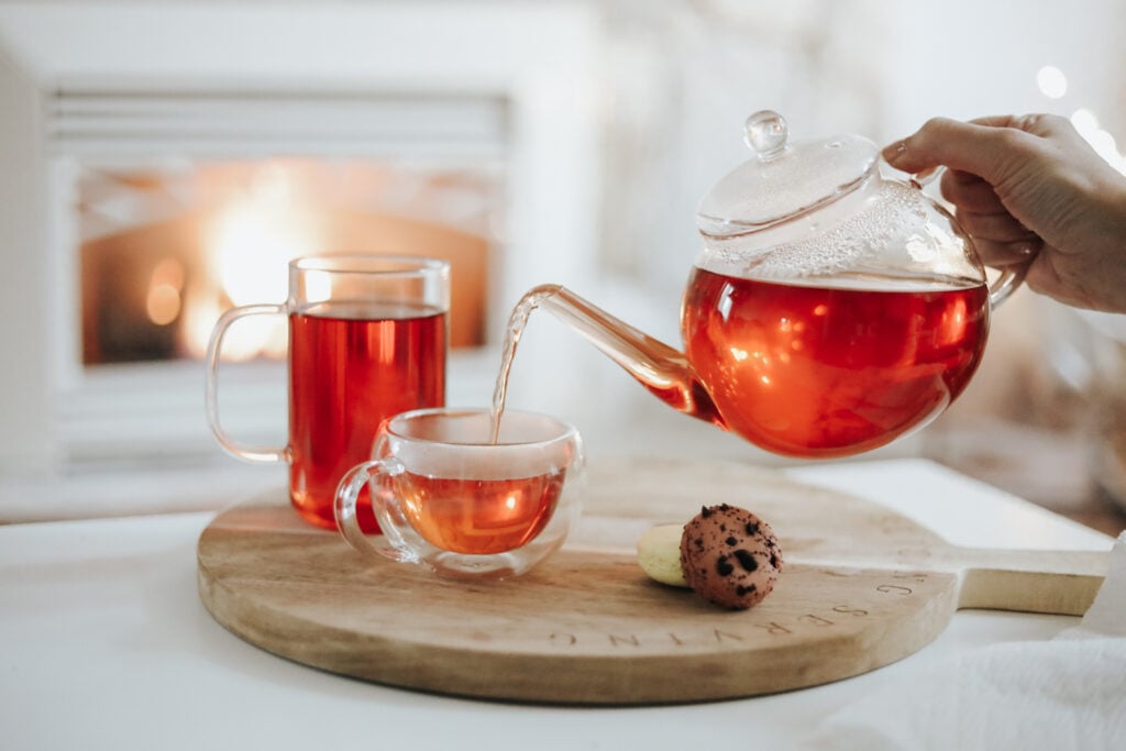 Hot tea in a teapot poured into two tea glasses in front of a fireplace.