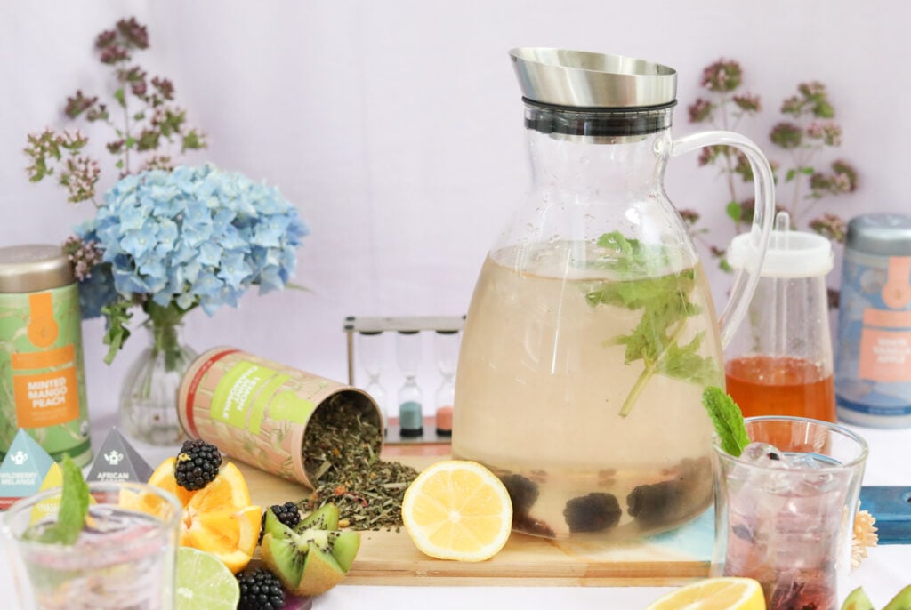 Iced tea pitcher with cut fruit, berries, and mint leaves with loose-leaf tea and fresh flowers.