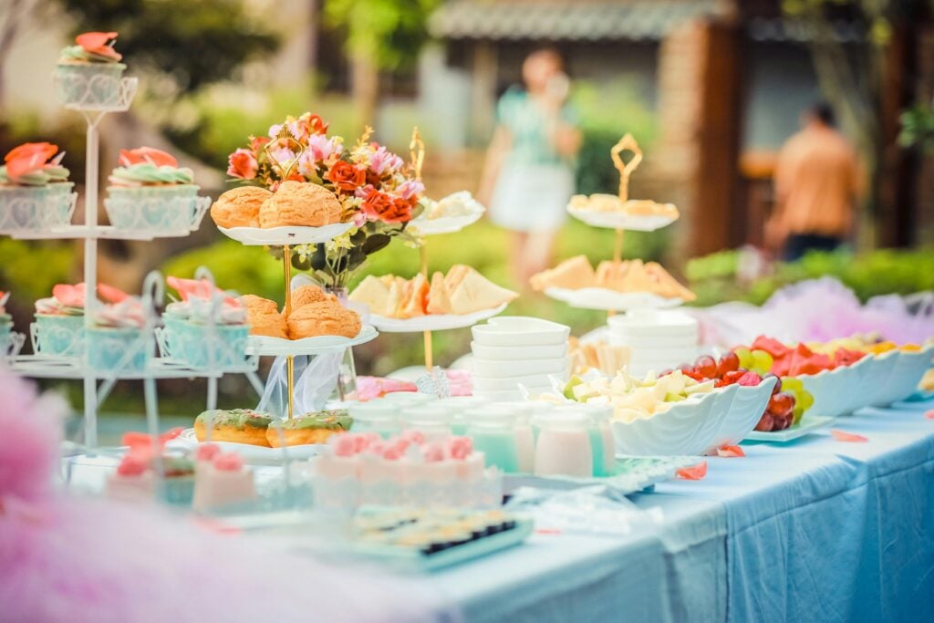 Outdoor afternoon tea party food table with pastries, sandwiches, and cut fruit.