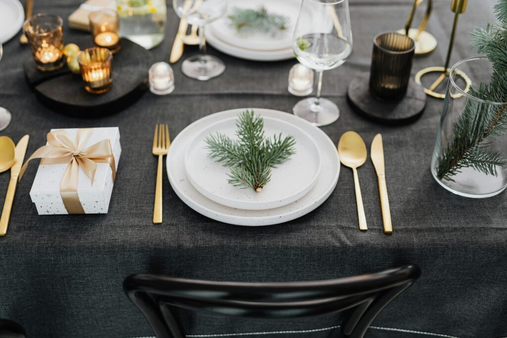 Elegant holiday tea party table setup with small gifts, gold silverware, white plates, candles, and pine tree fronds.