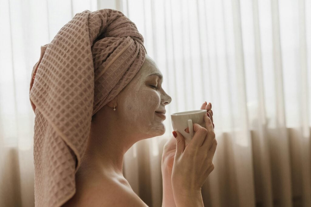 A woman with a beige hair towel sipping on a cup of tea while enjoying a face mask with tea for skin.