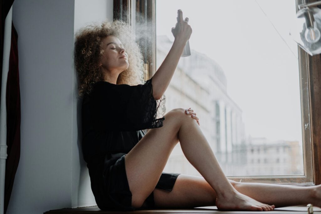 A woman with curly hair sitting by a window with a black robe and spritzing herself with a tea toner for skin.