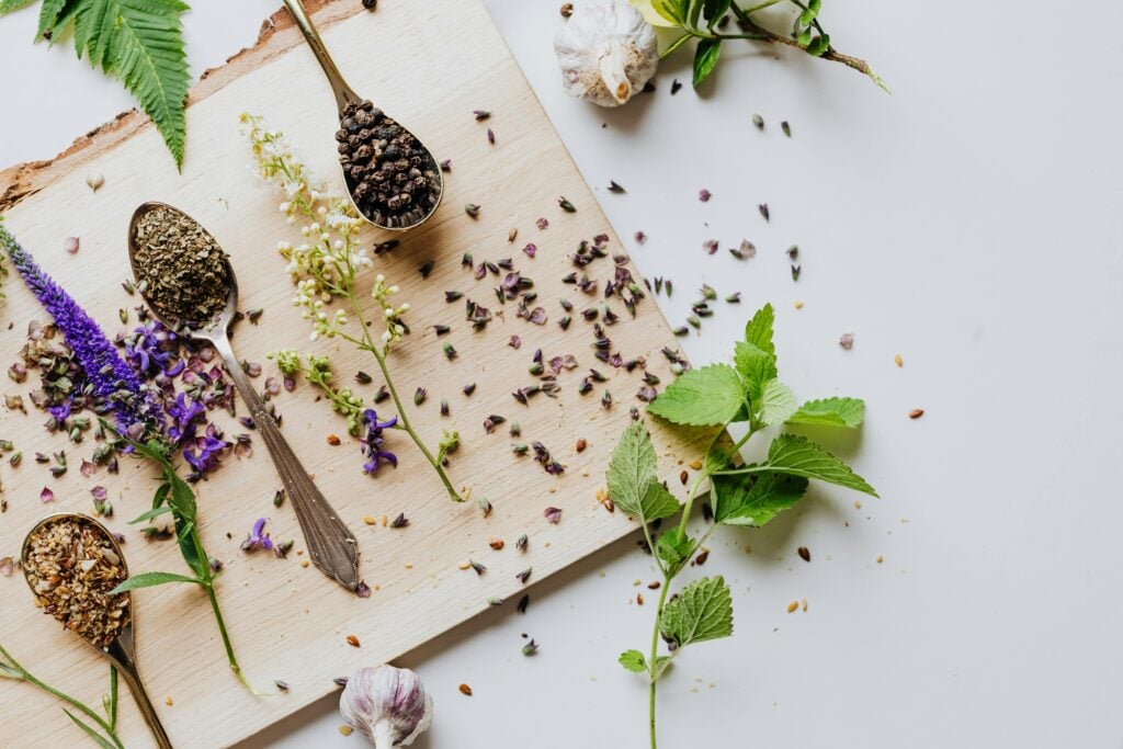 Silver spoons on a wood cutting board filled with spices surrounded by fresh herbs and flowers.
