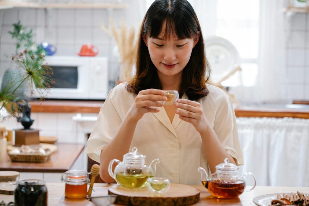 A woman with brown hair drinking holding a small glass teacup and steeping immune boosting teas in glass teapots.