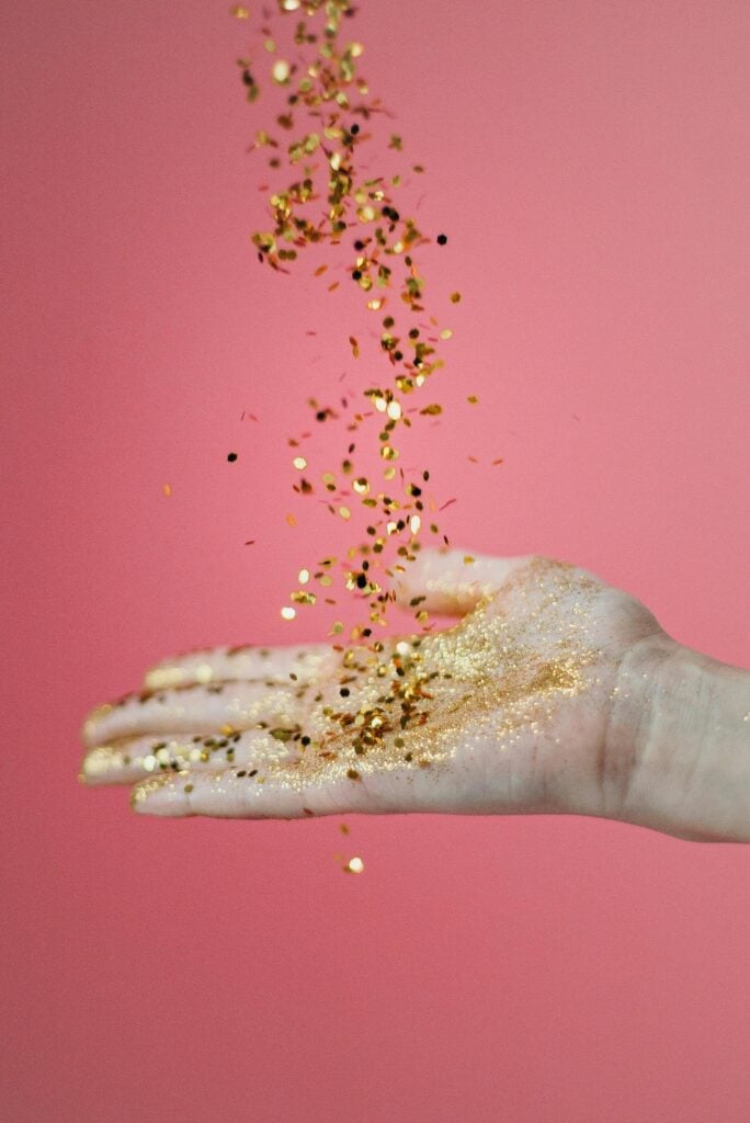 A hand with gold glitter pouring into it against a bright pink background.