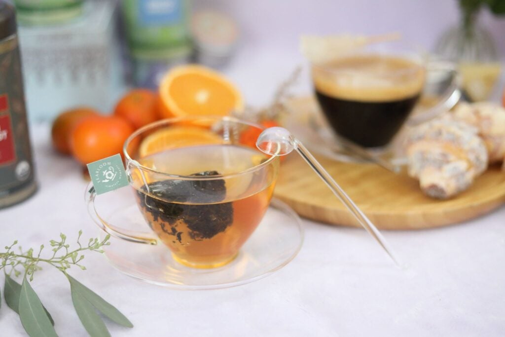 A classic shaped glass teacup with a small glass teaspoon on a table with a coffee cup and fresh fruit.