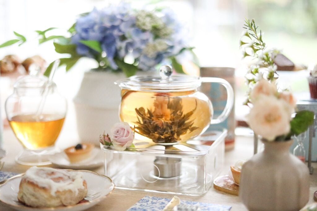 An eastern-style glass teapot with a blooming tea flower on a warming pedestal surrounded by tea treats, accessories, and flowers.
