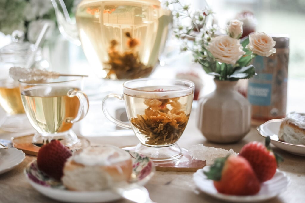 Footed glass teacups with blooming tea on a table with a glass teapot, pastries, strawberries, and fresh flowers.